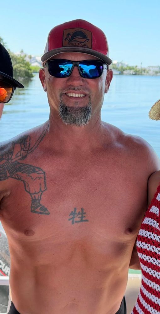 Smiling man wearing sunglasses and a cap, standing shirtless by a waterfront.