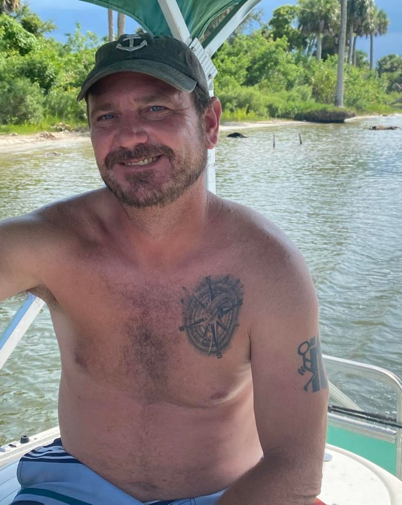 Man sitting shirtless on a boat with tattoos, smiling.